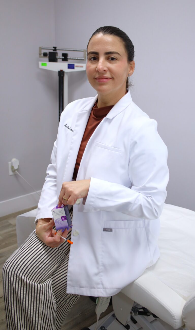 A female medical professional in a white lab coat sitting in a clinical exam room, holding a box of Botox Cosmetic and a medical syringe.