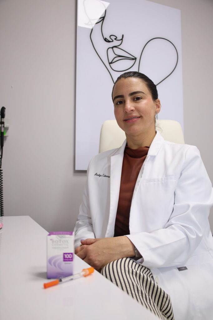 A female medical professional with dark hair tied back, wearing a white lab coat over a rust-colored top, sitting at a white desk in a clinical office. A box of Botox Cosmetic and a medical syringe sit on the desk in the foreground, while a minimalist line-art portrait of a woman's face hangs on the wall behind her.