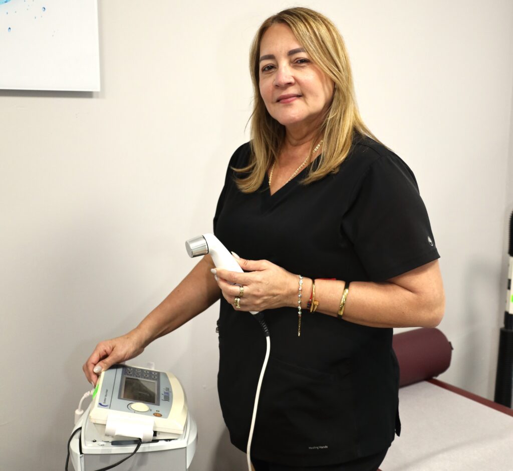 A female medical professional with blonde-highlighted hair, wearing black scrubs and gold jewelry, standing in a clinical room. She holds an ultrasound transducer in her right hand and rests her left hand on a control unit, looking towards the camera with a gentle expression.